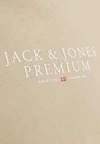 Bežo medvilnės audinio marškinėliai su iškiliais baltais užrašais "JACK & JONES PREMIUM" ir maža raudona Danijos vėliavos detalė.