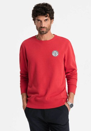 Sweatshirt rouge en mélange de coton, col rond, poignets et ourlet côtelés. Présente un logo circulaire sur le côté gauche de la poitrine. Porté avec un pantalon bleu marine.