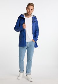 Blauwe Waterdichte Jas met Een Kap, Rits Voren en Drukknoopsluitingen. Heeft Een Grijze Voering, Twee Voorkantzakken en Een Relaxte Pasvorm. Gecombineerd met Lichtblauwe Jeans.