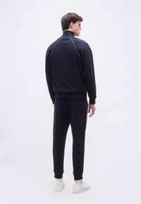 Άνδρας που στέκεται με την πλάτη στραμμένη, φορώντας μπλε ναυτικό φόρμα με ανοιχτόχρωμες λεπτομέρειες και λευκά αθλητικά παπούτσια, σε απλό λευκό φόντο.