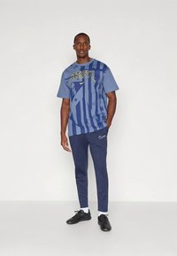 Blå grafik t-shirt med gule bogstaver, striber og stjernemønstre, kombineret med marineblå bukser og sorte sportssko.