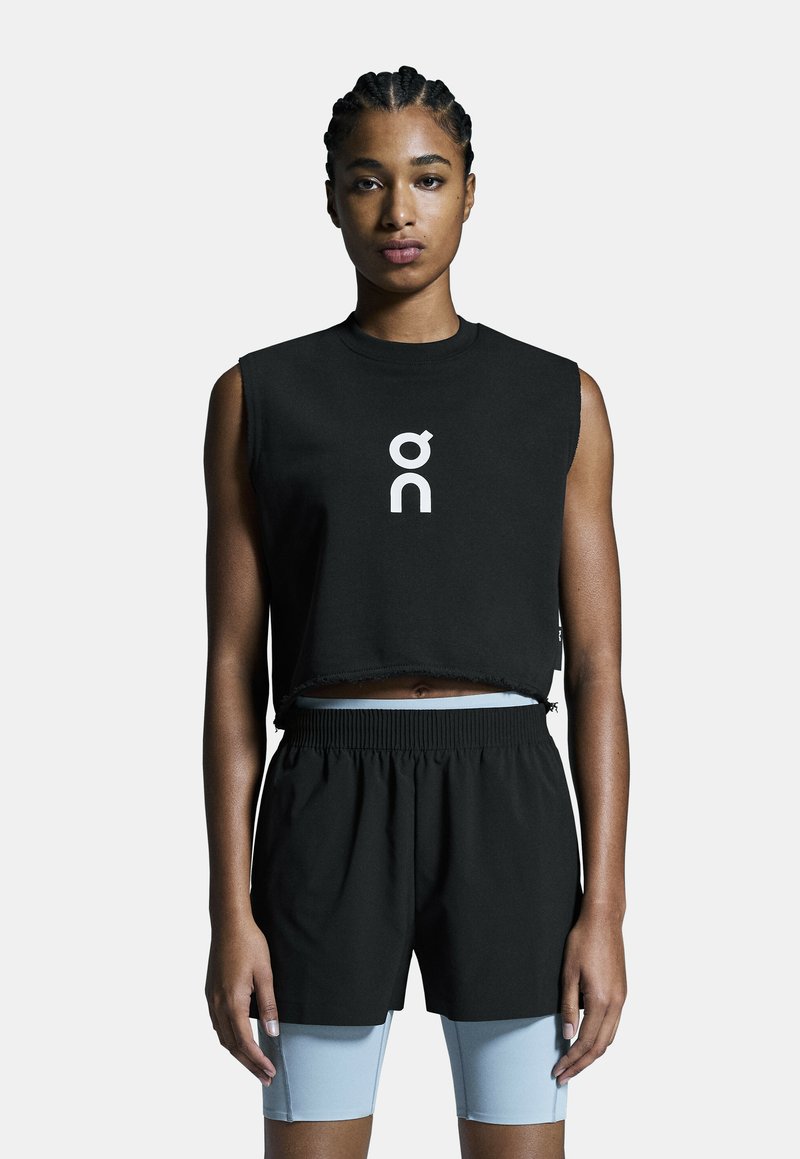 Schwarzes, kurz geschnittenes, ärmelloses Oberteil mit weißem Logo und fransigem Saum, kombiniert mit schwarzen Shorts und darunter hellblauen Fahrradhosen.