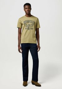 Oliivinvihreä puuvillapaita, jossa on musta grafiikka ”WRANGLER 1947”, yhdistettynä tummansinihin farkkuihin ja ruskeisiin nilkkapaitoihin.