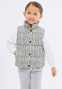 Zateplená vesta s čierno-bielym kvetinovým vzorom, vysokým golierom, patentkami a dvoma bočnými vreckami, nosená cez bielu košeľu s dlhým rukávom.