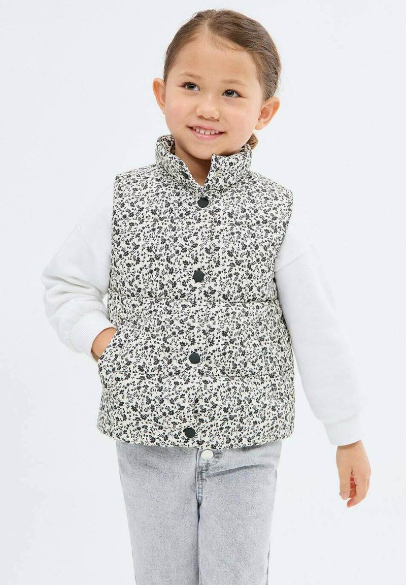 Zateplená vesta s čierno-bielym kvetinovým vzorom, vysokým golierom, patentkami a dvoma bočnými vreckami, nosená cez bielu košeľu s dlhým rukávom.