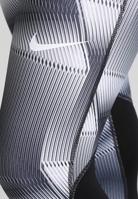 Czarno-białe legginsy z geometrycznym wzorem liniowym. Posiadają gładką teksturę, logo Nike oraz czarne przeszycia wzdłuż szwu.