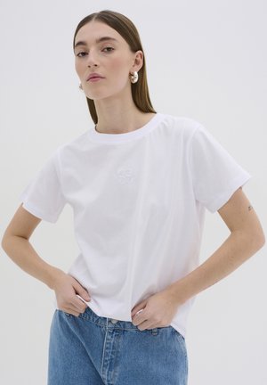 T-shirt bianco in cotone con maniche corte, scollo rotondo, con logo ricamato sul petto. Fit casual, abbinato a jeans blu.