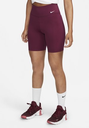 Personne portant un short cycliste Nike bordeaux, des chaussettes blanches Nike et des chaussures de course Nike bordeaux, debout devant un fond blanc uni.