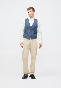 Μπλε γιλέκο με λεία υφή και ασημένια κουμπιά, φορεμένο πάνω από λευκό πουκάμισο και συνδυασμένο με μπεζ παν trousers και μαύρα παπούτσια.