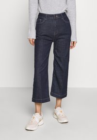 Wide-leg dark denim jeans with visible stitching and front pockets, paired with light-colored sneakers and a gray long-sleeve sweater.