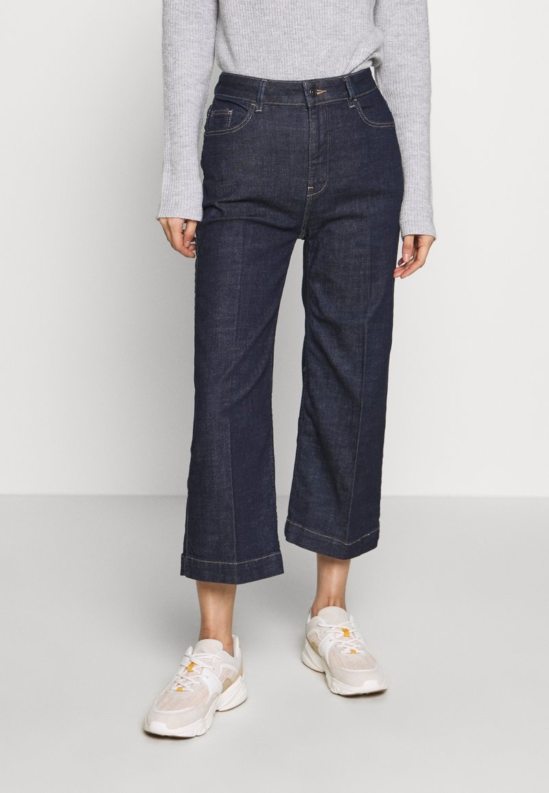 Wide-leg dark denim jeans with visible stitching and front pockets, paired with light-colored sneakers and a gray long-sleeve sweater.