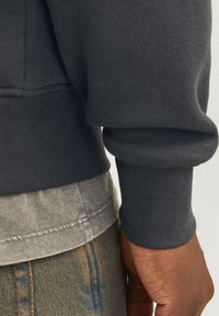 Ciemnoszara bluza z ściągaczami, noszona na jasnoszarym T-shircie. Widoczne szwy na dole i dżinsowy materiał w talii.