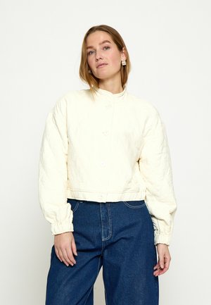 Žena so svetlo hnedými vlasmi nosí krémovú prešívanú bundu na gombíky s elastickými manžetami a tmavomodré džíny s vysokým pásom na jednoliatom pozadí.