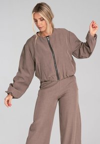 Czekoladowa, krótka kurtka bomberka z zamkiem z przodu, luźnymi rękawami i ściągaczem na dole. Noszona z dopasowanymi spodniami typu wide-leg. Tekstura wydaje się gładka.