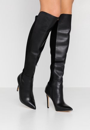 Czarne skórzane kozaki za kolano z ostrymi czubkami i obcasami typu szpilka, noszone na widocznych dolnych częściach nóg na jednolitym tle.