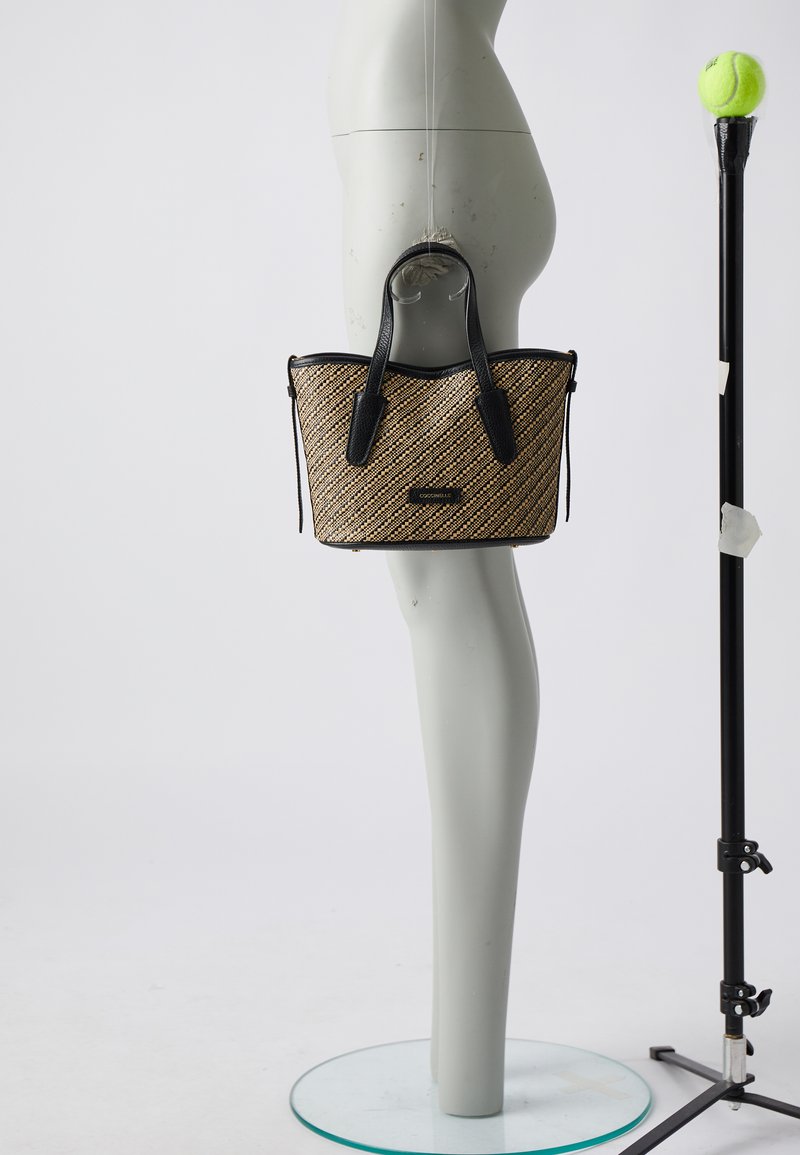 Woven straw tote bag in tan with black leather accents. Features dual handles, an open top, and a small logo plate on the front.