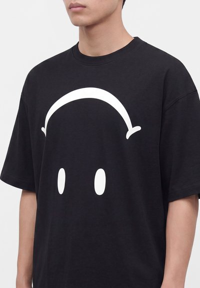 Homme portant un t-shirt noir avec un grand dessin minimaliste de visage blanc montrant deux yeux ovales et une ligne arquée au-dessus.