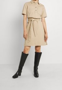 Mannequin portant une robe beige ceinturée à manches courtes et des bottes noires montantes à talons, debout devant un fond clair uni.