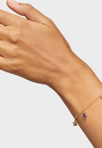 Bracciale in oro con due gemme pendenti: una ovale di un profondo blu e una rotonda di un rosa chiaro, posizionato su un polso abbronzato.