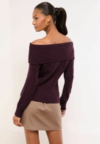 Olkapäät paljastava, tummanvioletti neulepaita, jossa on letitetyt kuvioinnit ja istuva malli, yhdistetty ruskeaan ruudulliseen hameeseen.