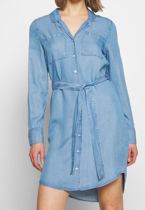 Frau, die ein hellblaues Jeanshemdkleid mit langen Ärmeln, Brusttaschen, Knopfleiste und einem gebundenen Taillengürtel trägt.
