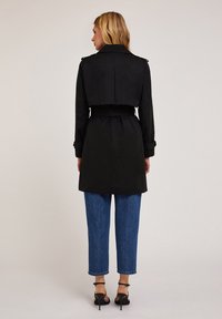 Palton negru de tip trench cu un design scurtat, talie evidențiată cu un brâu și epoleți pe umeri, asociat cu blugi albaștri cu talie înaltă și tocuri negre.