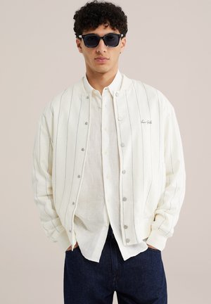 Mladý muž nosiaci tmavé slnečné okuliare, biely saka s tenkými pásikmi, bielu košeľu na gombíky a tmavé džínsy, stojaci s rukami v vreckách.