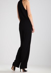 Combinaison noire sans manches avec une taille cintrée, conception à jambes larges et tissu lisse. Associée à des talons hauts noirs.