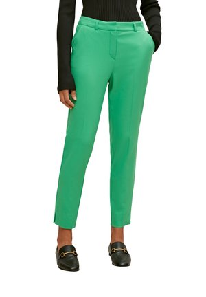 Persoon draagt felgroene, op maat gemaakte broek die tot de enkels reikt, een zwart geribd top met lange mouwen en zwarte loafers met gouden accenten.