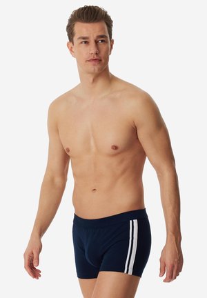 Man zonder shirt die naar links kijkt, gekleed in marineblauwe boxershorts met twee witte verticale strepen aan de zijkant, staand tegen een effen witte achtergrond.