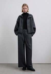 Chaqueta extragrande de color gris oscuro con cuello, combinada con pantalones de pierna ancha y un cuello alto negro ajustado. Tejido liso y corte holgado.