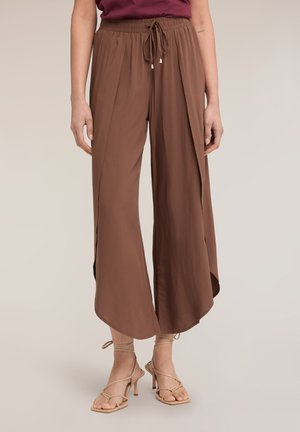 Vrouw draagt losse bruine wijde broek met zijsplitsingen en beige hooggehakte sandalen met bandjes, staand tegen een effen achtergrond.