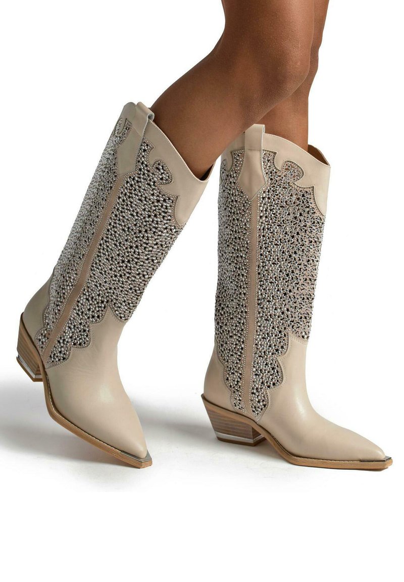 Stivali cowboy beige con punta appuntita, caratterizzati da un'incisione testurizzata in argento sul gambale e un tacco in legno leggermente inclinato.