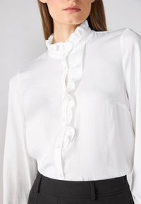 Blouse blanche à manches longues avec un col volanté et une fermeture à boutons, fabriquée en tissu léger ; assortie à un pantalon noir ajusté.