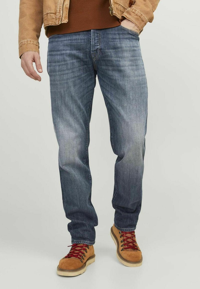 Jeans de mezclilla azul oscuro con un acabado desgastado, corte tapered, cinco bolsillos y un lazo para cinturón. Combinados con botas de senderismo color beige y cordones rojos.