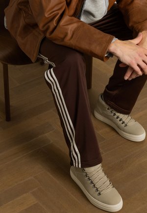 Bruine trainingsbroek met witte strepen aan de zijkanten, gecombineerd met beige hoge sneakers. De textuur lijkt glad te zijn, zittend op een houten stoel.