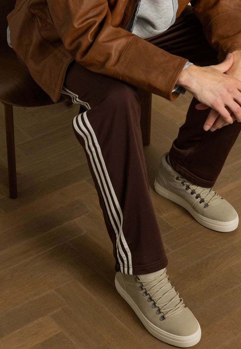Bruine trainingsbroek met witte strepen aan de zijkanten, gecombineerd met beige hoge sneakers. De textuur lijkt glad te zijn, zittend op een houten stoel.