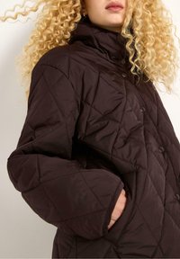 En person med krøllet blond hår har en mørkebrun, diamantquiltet jakke på, der fremhæver et nærbillede af jakkens krave og lomme.