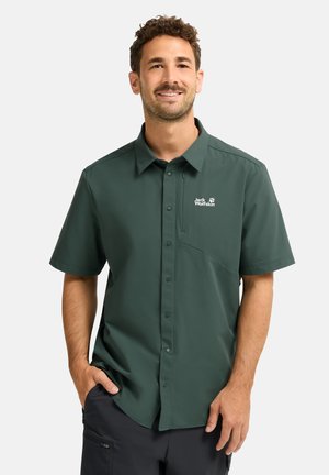 Uomo sorridente, indossa una camicia verde scuro a maniche corte con bottoni e logo "Jack Wolfskin" e pantaloni neri, con le mani in tasca.