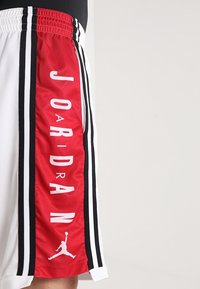 Shorts de basketball rouges et blancs avec des rayures noires sur les côtés. Présente un grand texte "JORDAN" et le logo emblématique du jumpman sur le côté. Tissu léger.