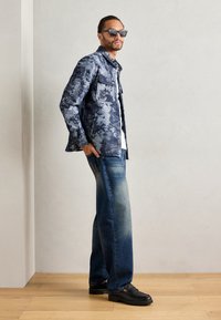 Blue patterned jacket with two chest pockets, white shirt underneath, faded blue jeans, and black loafers; person wearing dark sunglasses.