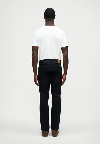 Άνδρας που στέκεται με την πλάτη του στραμμένη προς την κάμερα, φοράει άσπρο t-shirt, μαύρο παντελόνι και καφέ παπούτσια, μπροστά σε απλό φόντο.