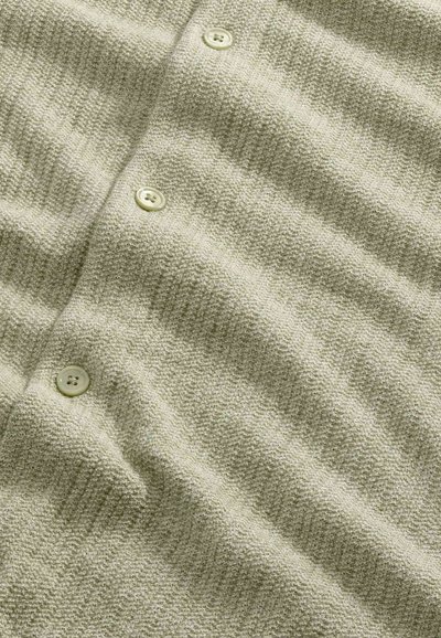 Tessuto a maglia beige chiaro testurizzato con tre piccoli bottoni rotondi disposti verticalmente su un indumento.