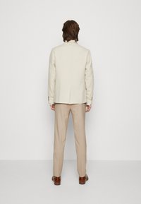 Beige linnekavaj med en enkel slits och matchande beiga byxor. Bruna läderskor kompletterar outfiten. Tyget verkar mjukt och strukturerat.