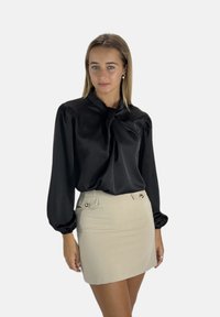 Blusa nera in satin con collo alto e maniche a palloncino abbinata a una mini gonna beige con bottoni decorativi sul davanti.
