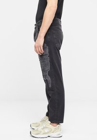Pantalones de mezclilla negros con un ajuste holgado, que presentan un parche desgastado en el muslo. Combinados con zapatillas de colores claros. Vista lateral.