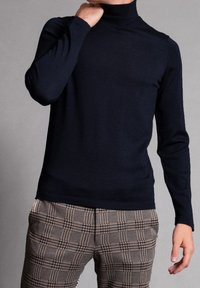 Navyfarbener Rollkragenpullover aus glattem Strickstoff, taillierte Passform mit langen Ärmeln. Getragen mit braun- und cremefarbenen karierter Hose.