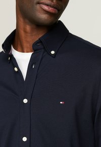 Camicia blu navy con bottoni, caratterizzata da un tessuto texturizzato, colletto a punta, bottoni bianchi e una piccola toppa con logo rosso, bianco e blu sul petto.