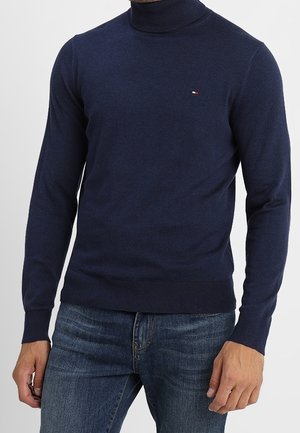 Mann, der einen eng anliegenden dunkelblauen Rollkragenpullover und blaue Jeans trägt, steht mit entspannten Händen an den Seiten.