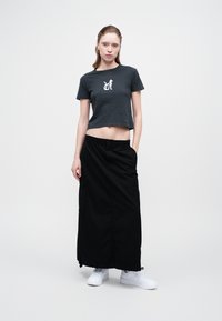 Žena nosi crni skraćeni T-shirt s grafikom bijelog mačka, dugu crnu suknju s džepovima i bijele tenisice, stoji s jednom rukom u džepu.